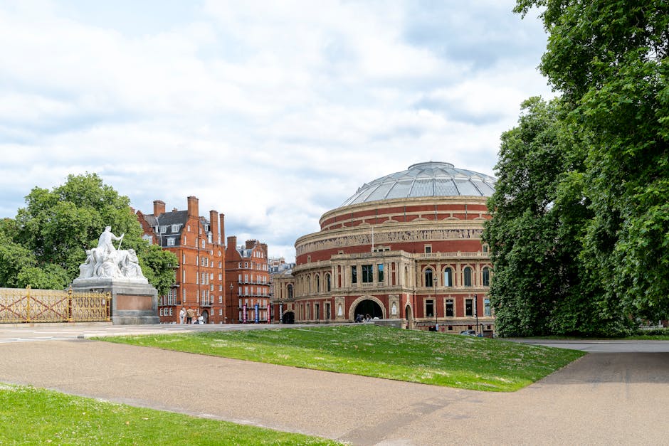 The Royal Albert Hall in London, a large circular building with a domed roof. The exterior features a red brick facade with decorative stone details and numerous arched windows. The front entrance has
