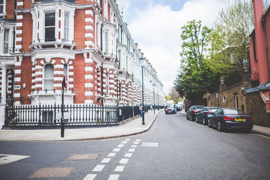 A residential street near Earls Court Station featuring a row of Victorian-style terraced houses with red and white brick facades, ornate architectural detailing, and black wrought iron fences. The street has parked cars along the curb, with a few pedestrians visible in the distance. The road surface appears clean and well-maintained, with clear white dashed lines and tactile paving at the corner. Bright daylight illuminates the scene, highlighting the neatness and historic charm of the buildings. This street exemplifies typical London residential architecture and is part of the area serviced by Cleaners Earls Court for surface cleaning and domestic hygiene maintenance, aligning with the town's emphasis on thorough cleaning and sanitisation of urban living spaces.