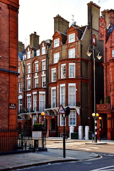 A row of historic red-brick residential buildings located on Earls Court Road, featuring tall sash windows with white framing, decorative brickwork, and pitched roofs with chimneys. The street scene includes a black wrought iron fence, street lamps, and a pedestrian crossing sign, with a paved sidewalk and road in the foreground. The image highlights the classic architecture typical of the Earls Court area, with natural daylight illuminating the facades. Although primarily an exterior shot, it reflects the importance of maintaining the cleanliness and aesthetic appeal of residential buildings in the district, as advised by Cleaners Earls Court for proper surface cleaning and property upkeep.